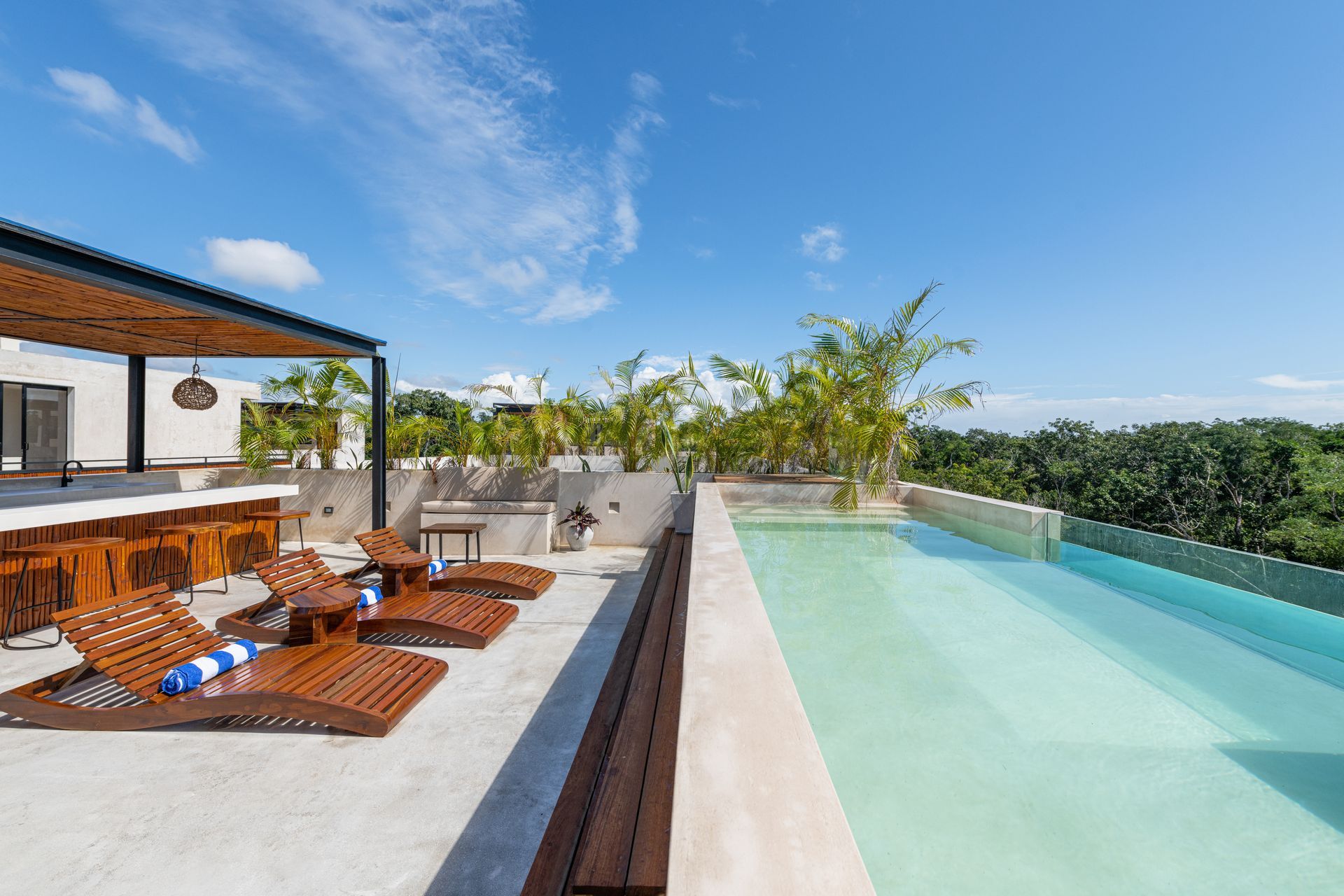 Rooftop infinity pool beside lounge chairs and a shaded terrace overlooking green hills and blue sky