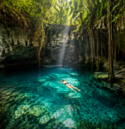 Sunlit turquoise cenote pool with a swimmer beneath hanging vines and rock walls