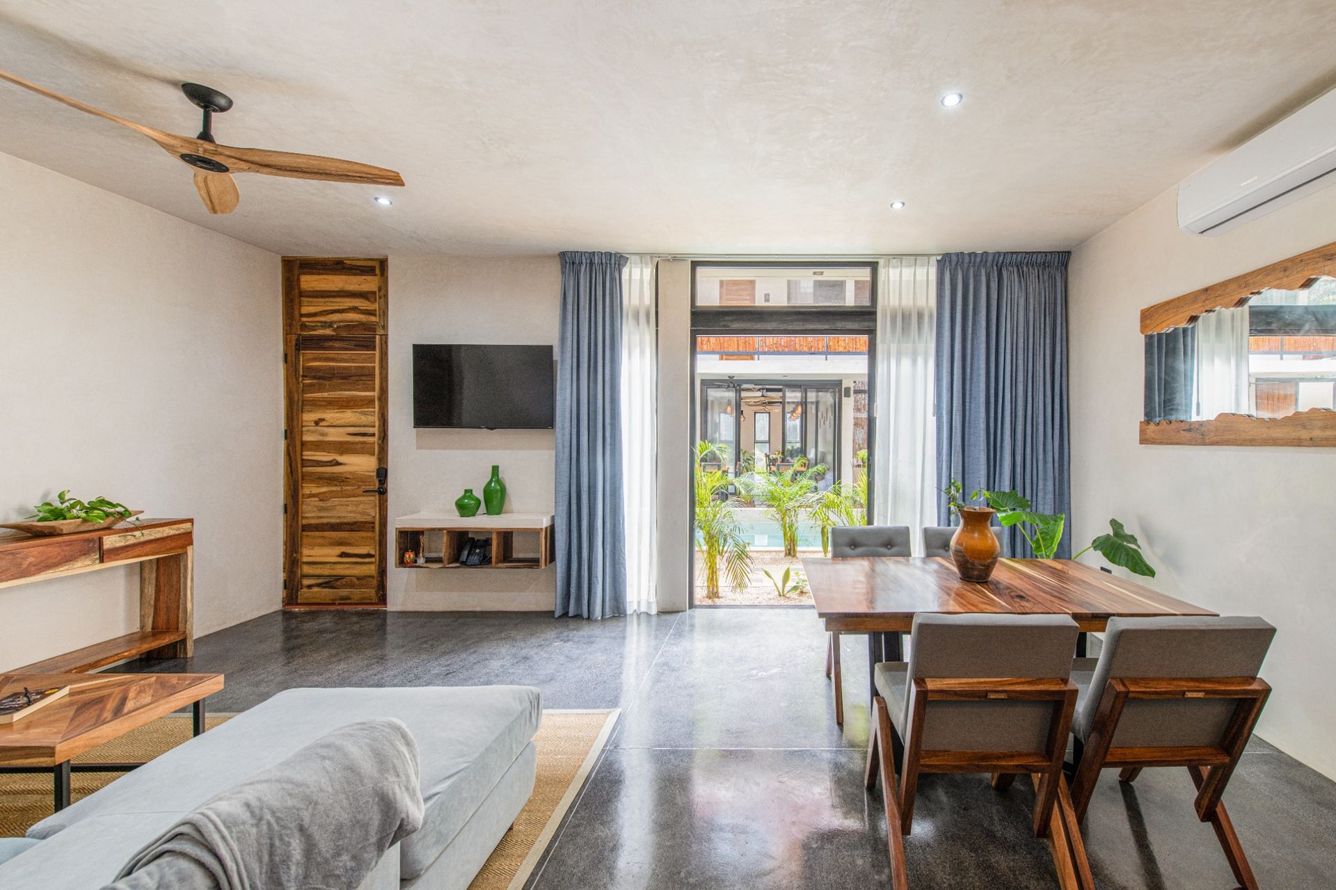 Bright living-dining room with sofa, TV, wooden table, plants, and sliding doors to a balcony