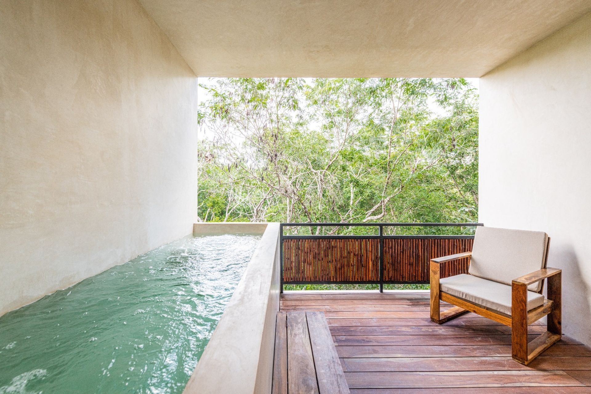 Indoor plunge pool beside a wooden deck with a bench, overlooking trees through an open wall