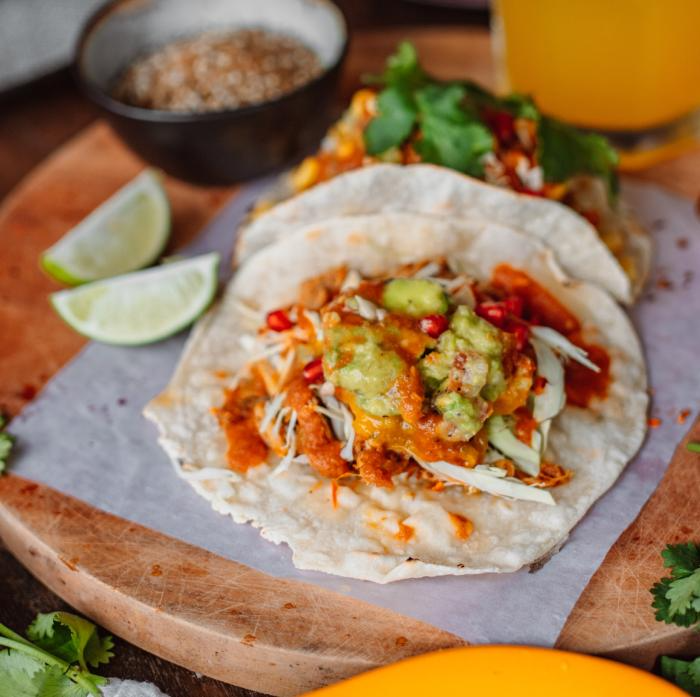 Tacos on a wooden board with guacamole, salsa, and cilantro, served with lime and a drink.