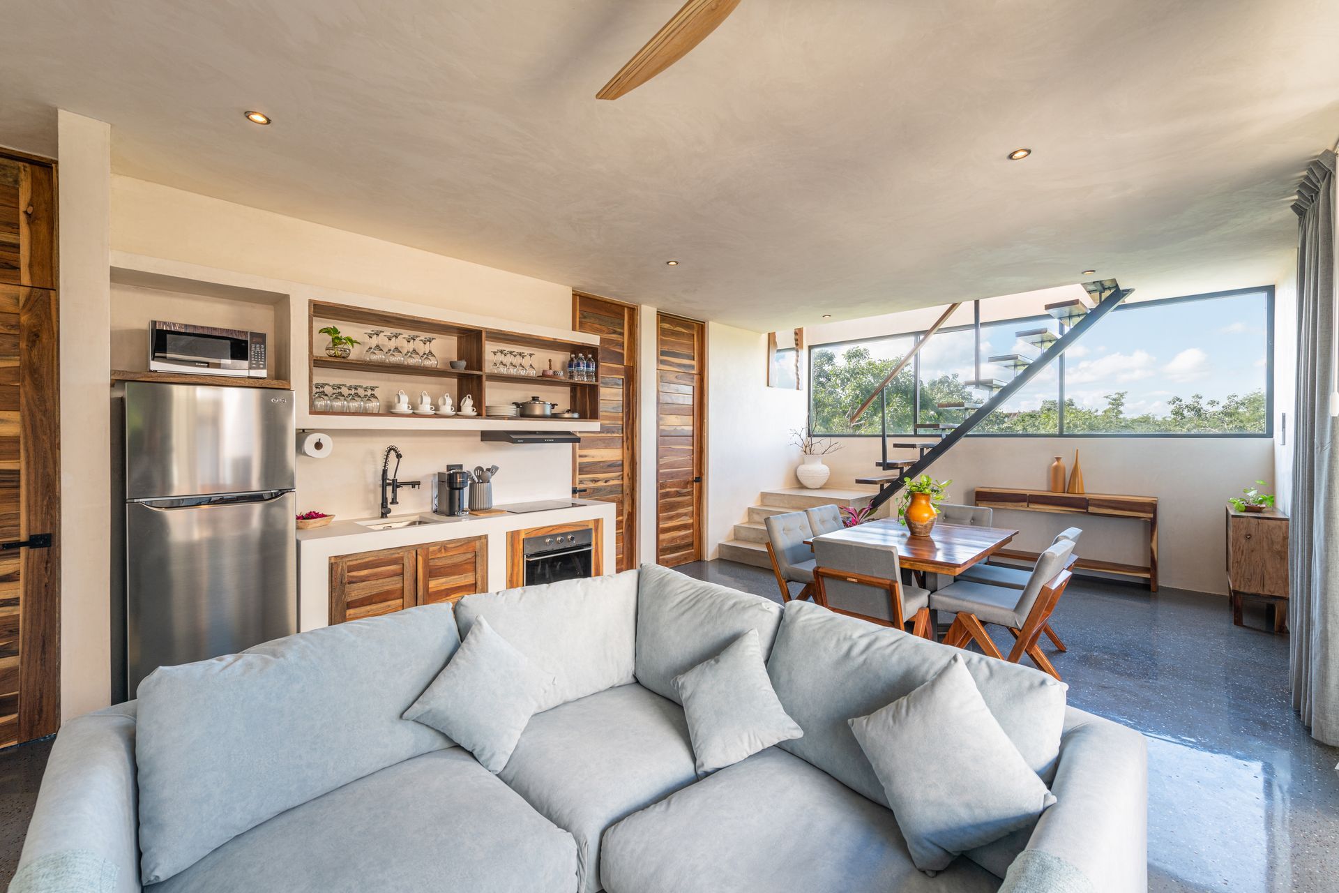Bright open-plan living room with gray sofa, kitchen, and dining area facing a balcony view