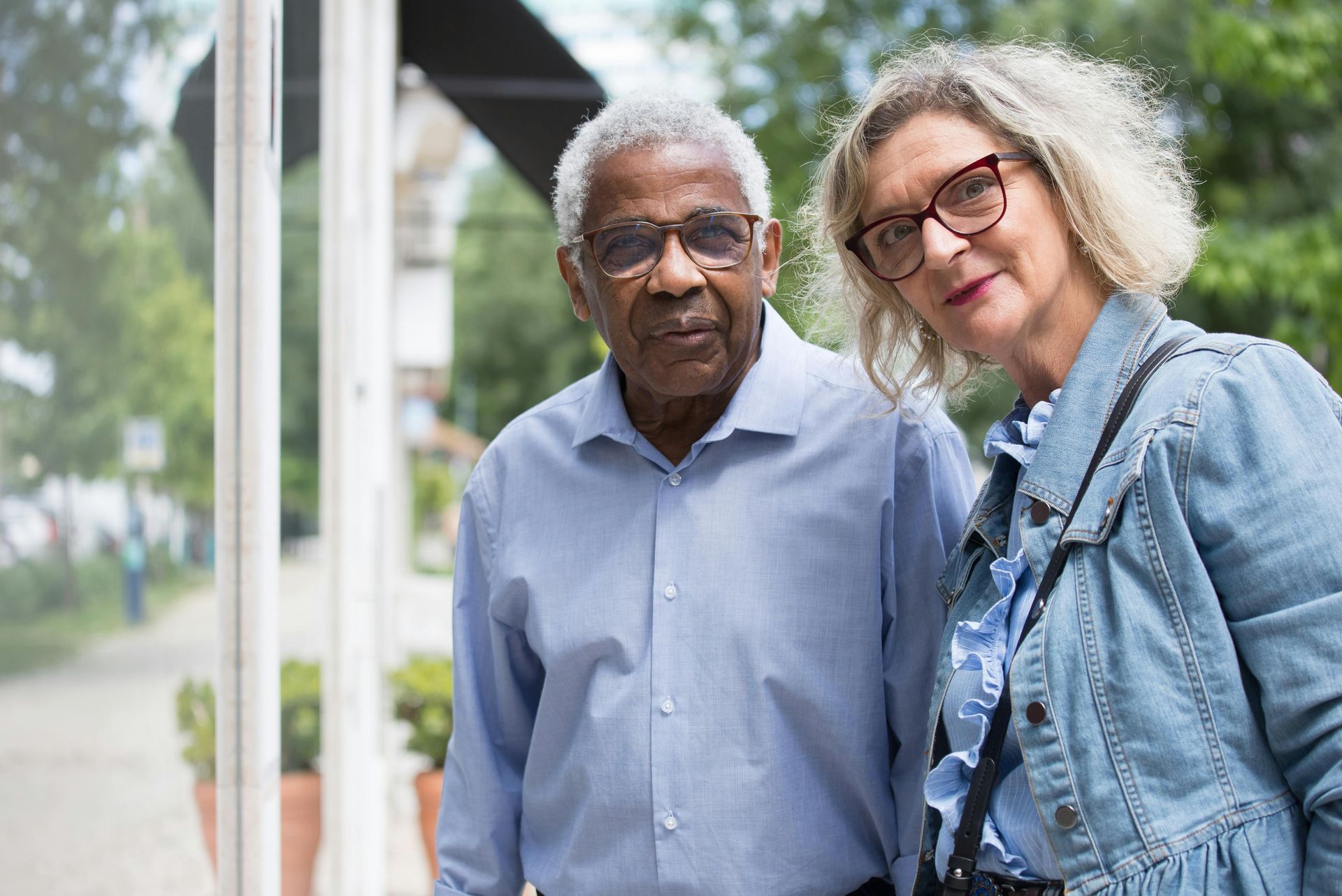 Two people wearing glasses stand side-by-side in an outdoor setting with trees and a walkway in the background.