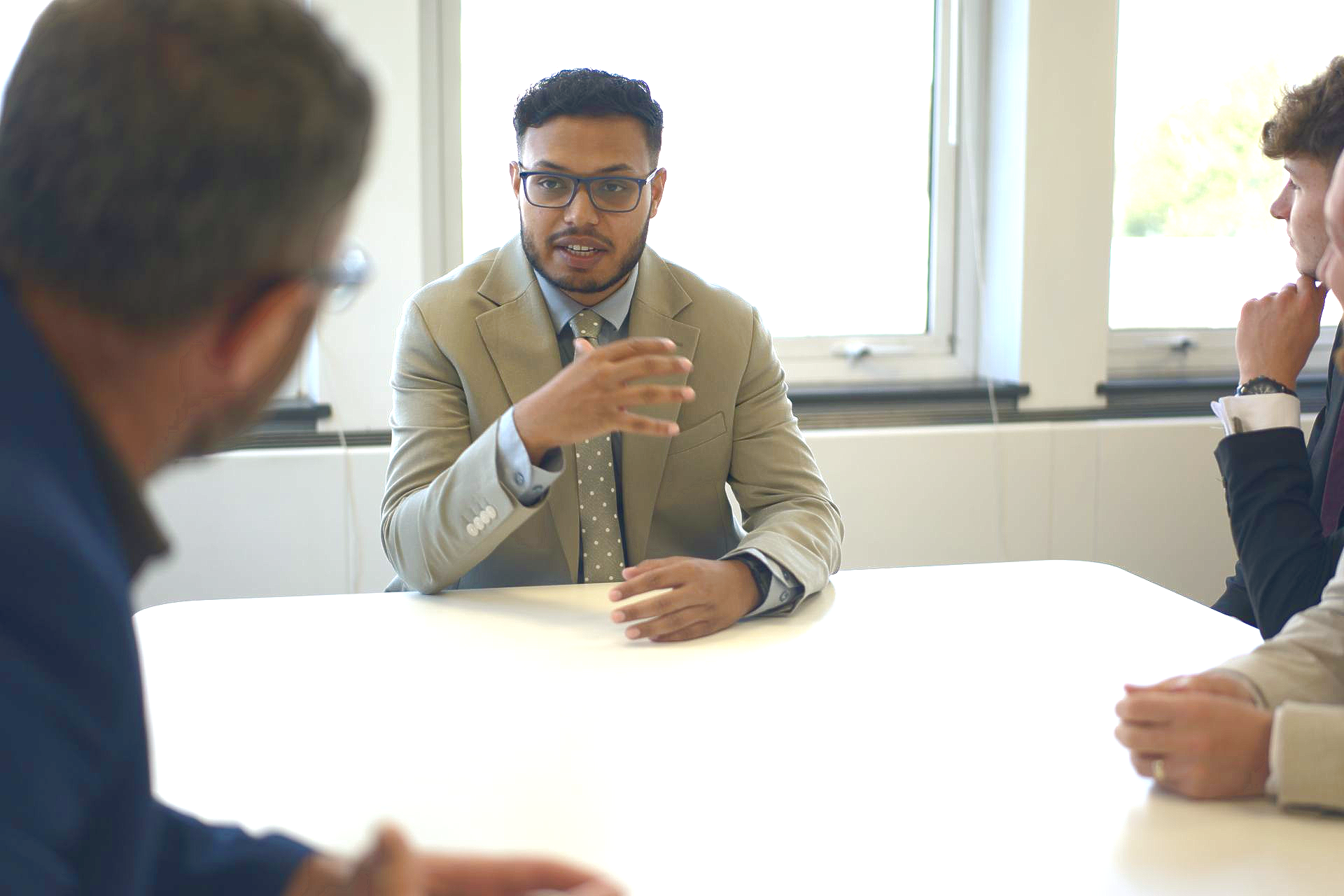 A group of people in business attire sit around a bright table, engaged in a discussion in a well-lit office.