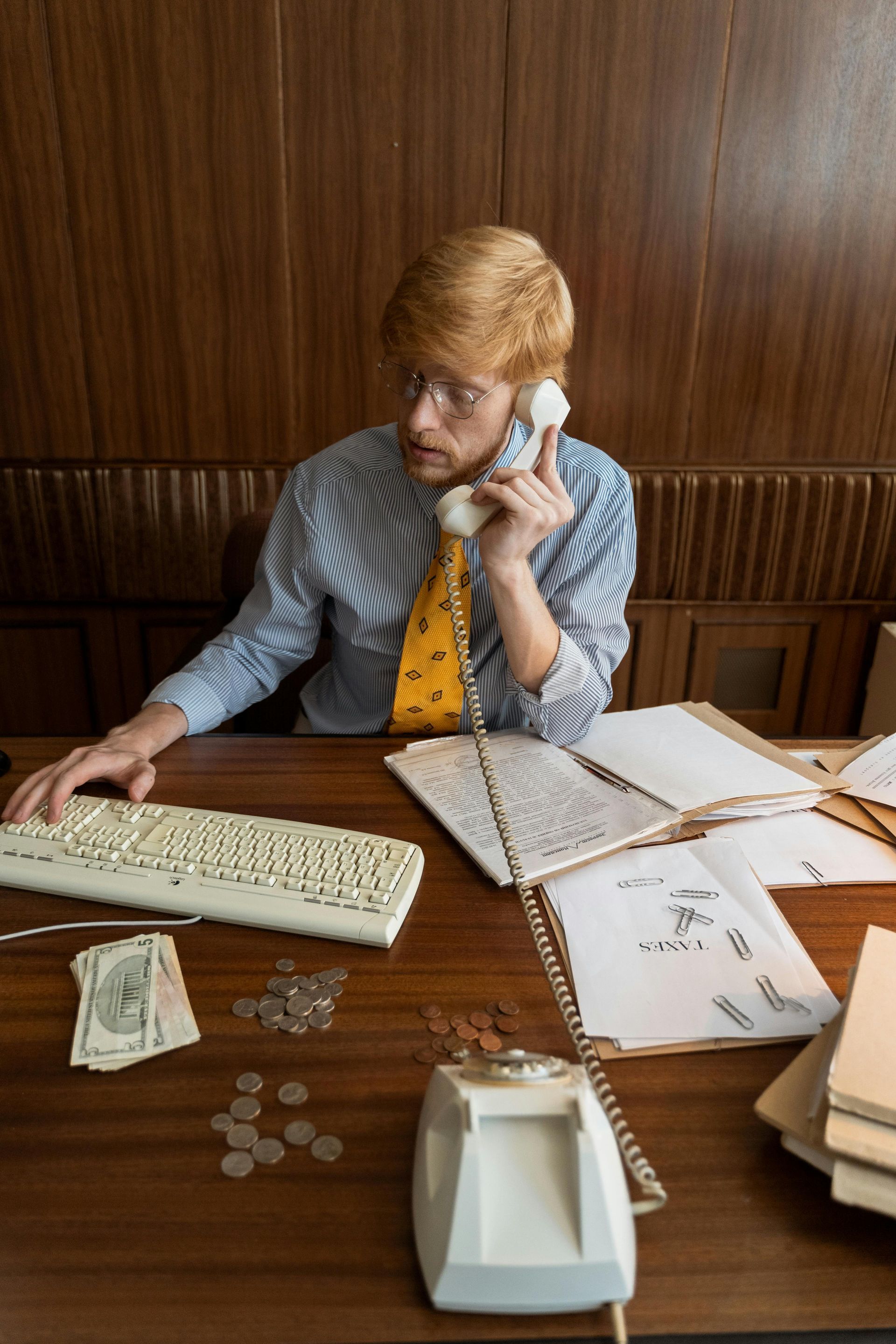Person at a desk in a wood-paneled office, holding a phone and looking at papers and a keyboard.