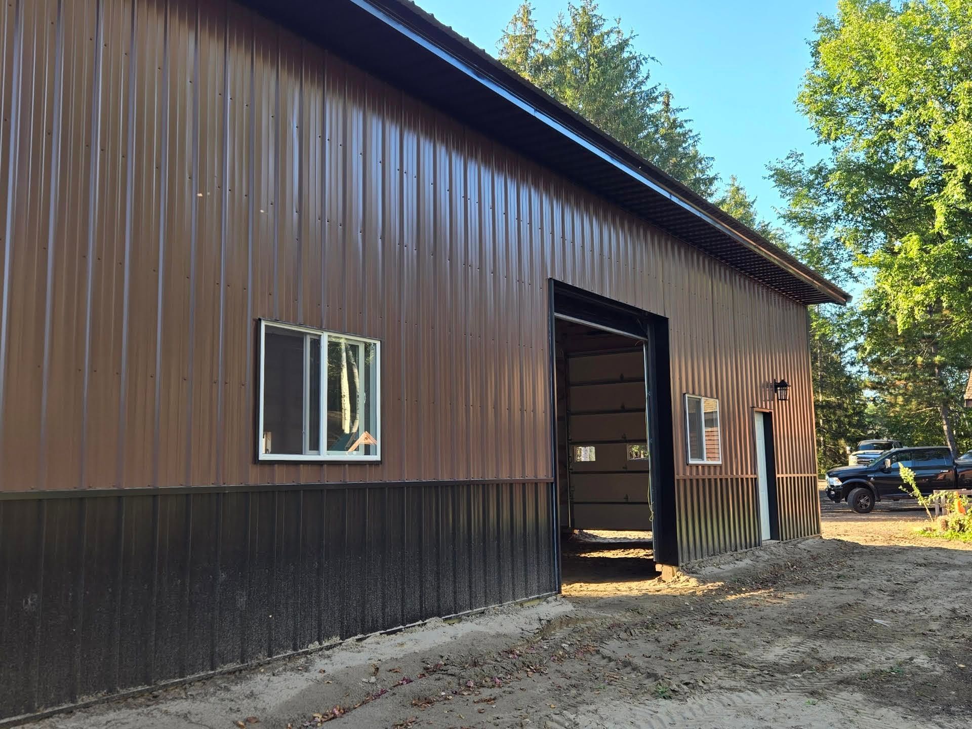 Brown barn-like building with a metal exterior, open doorway, and gravel ground beside trees