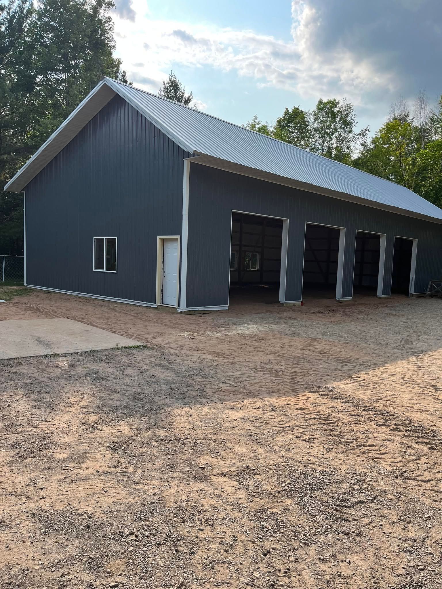 Gray metal barn with a wavy roof and open bays on a gravel lot under a cloudy sky