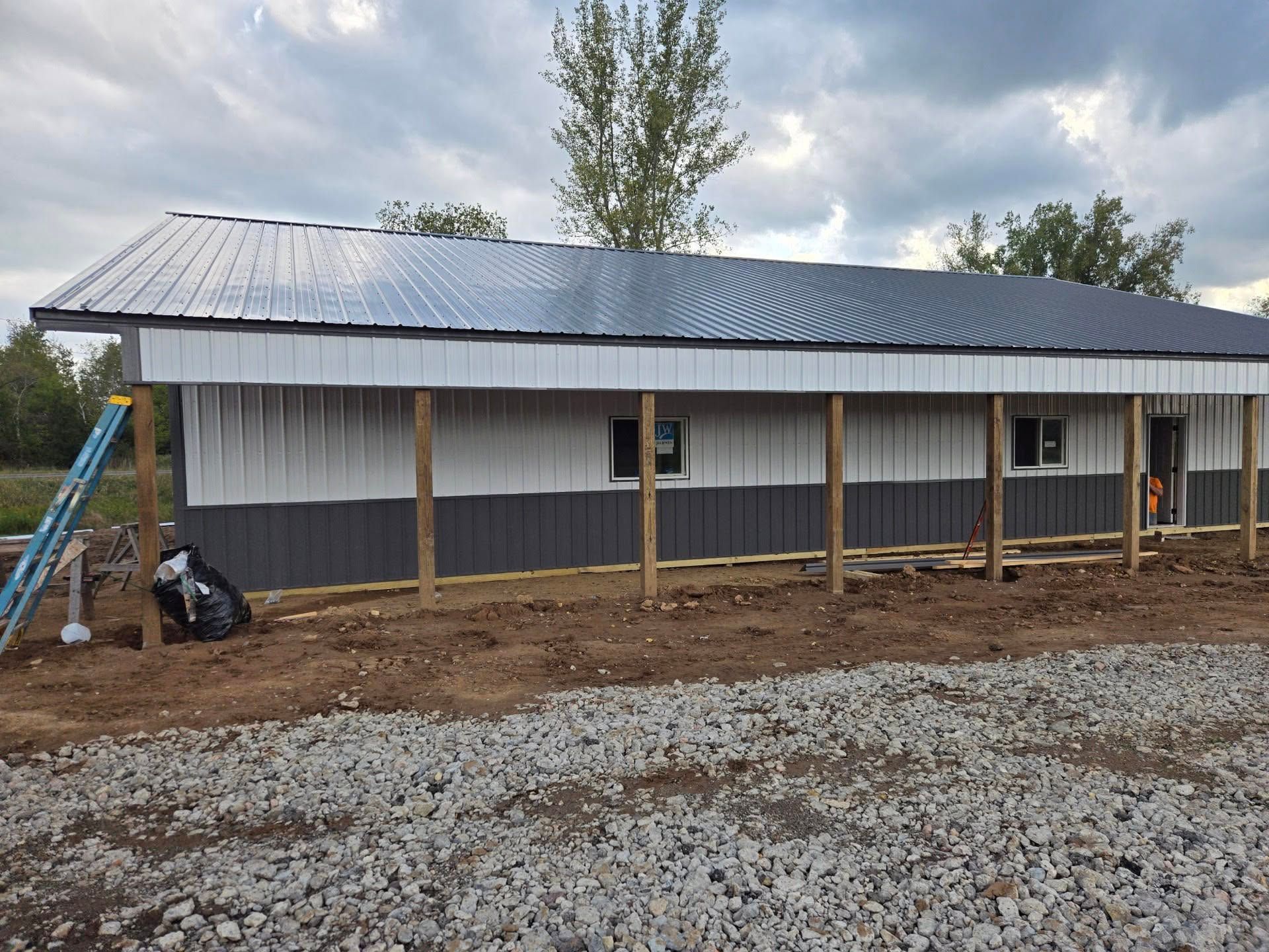 Long metal-roofed building under construction with wooden posts and gravel foreground