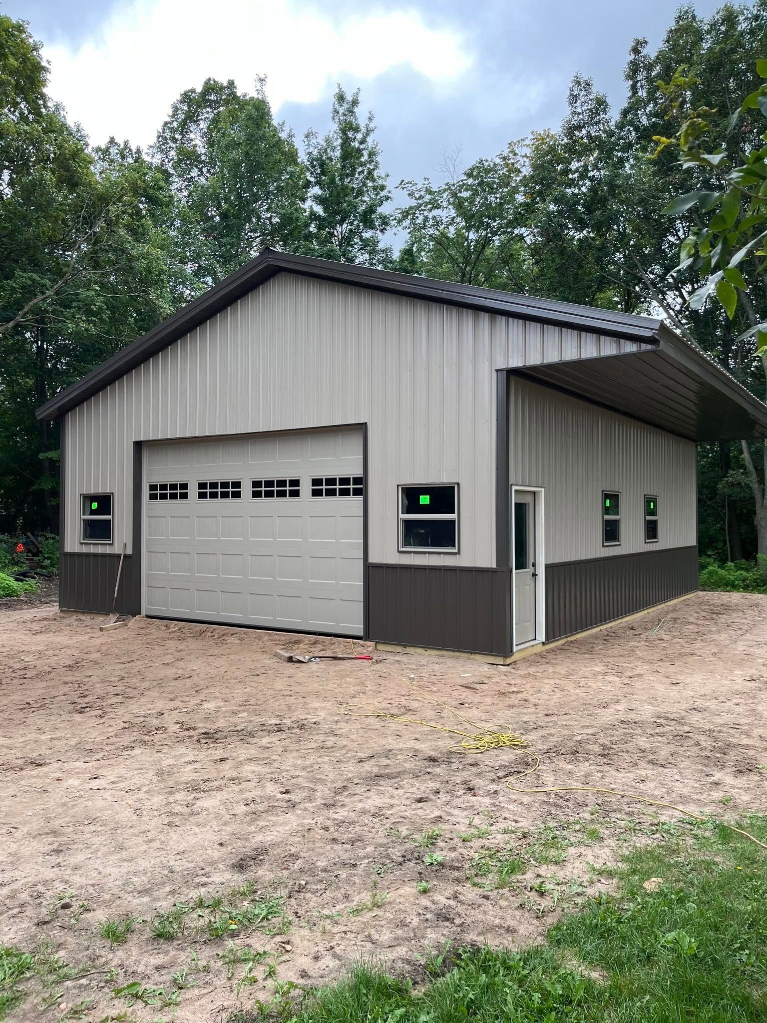 Small gray metal garage building with a white overhead door, set on a gravel lot amid trees.