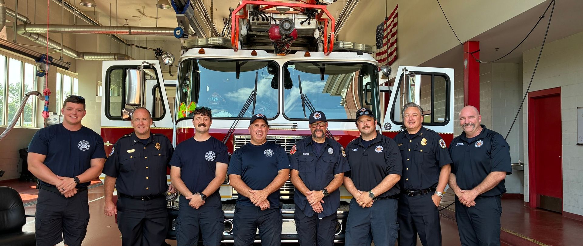 Firefighters stand in front of a fire truck in a fire station.