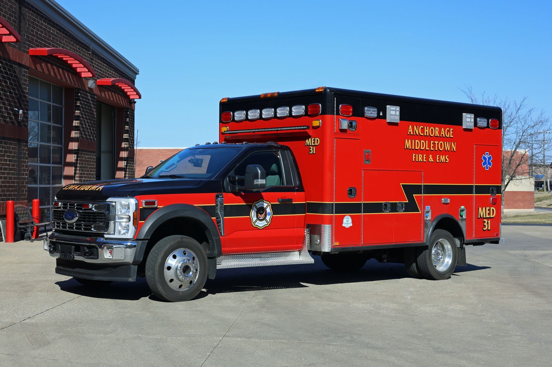 Red and black ambulance parked in front of a brick building on a sunny day.