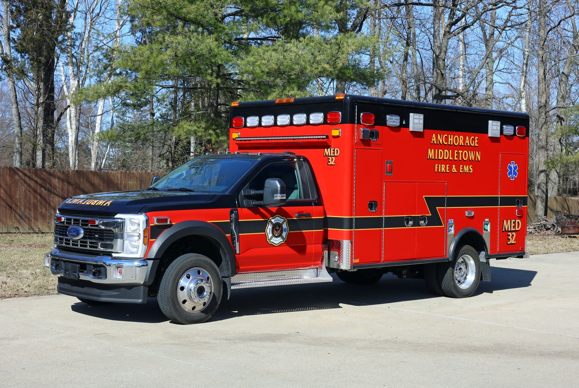 Red and black ambulance truck parked on asphalt road, with trees in the background.