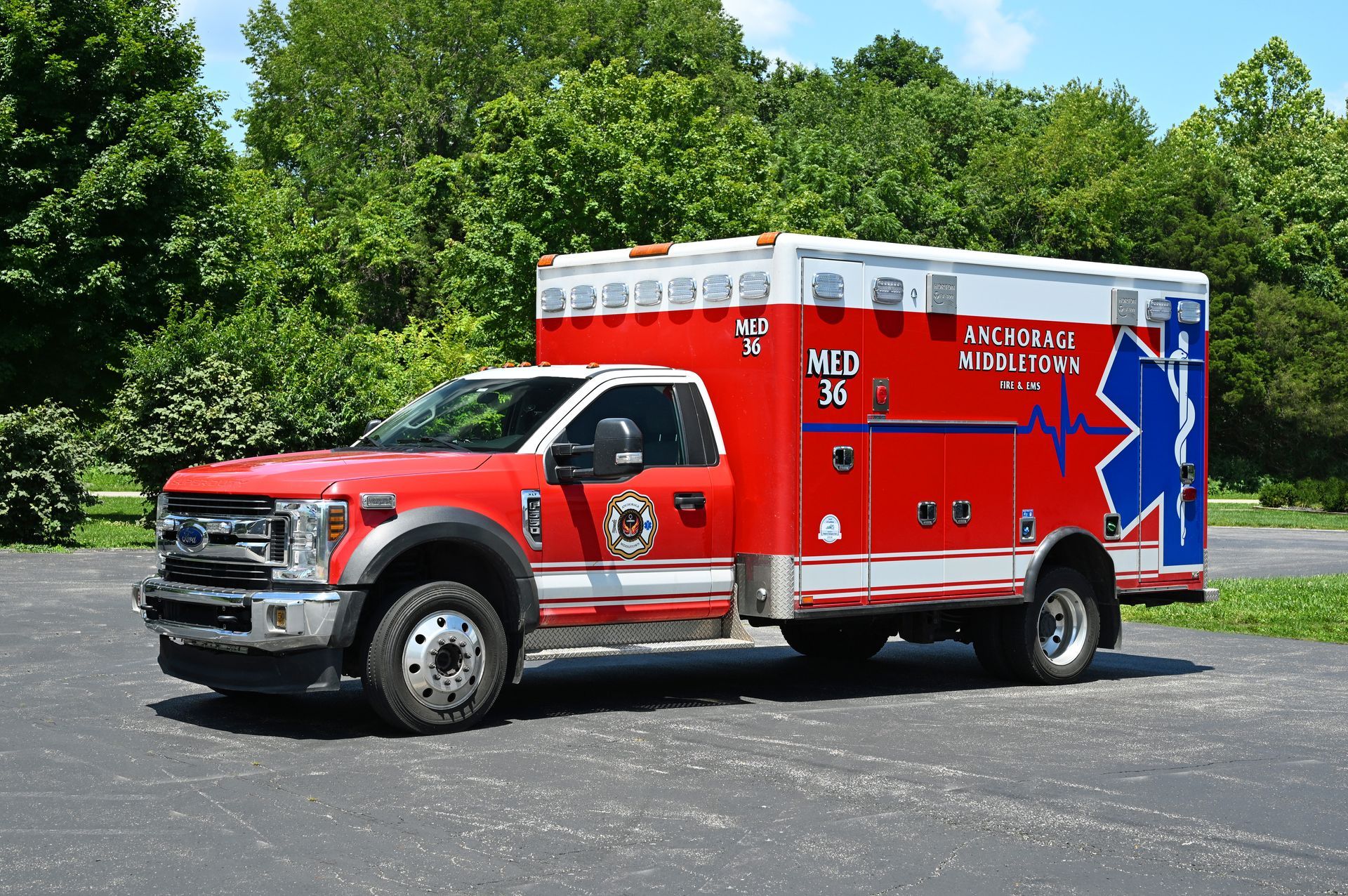 Red and white ambulance truck with a blue Star of Life emblem, parked on pavement, with trees in background.