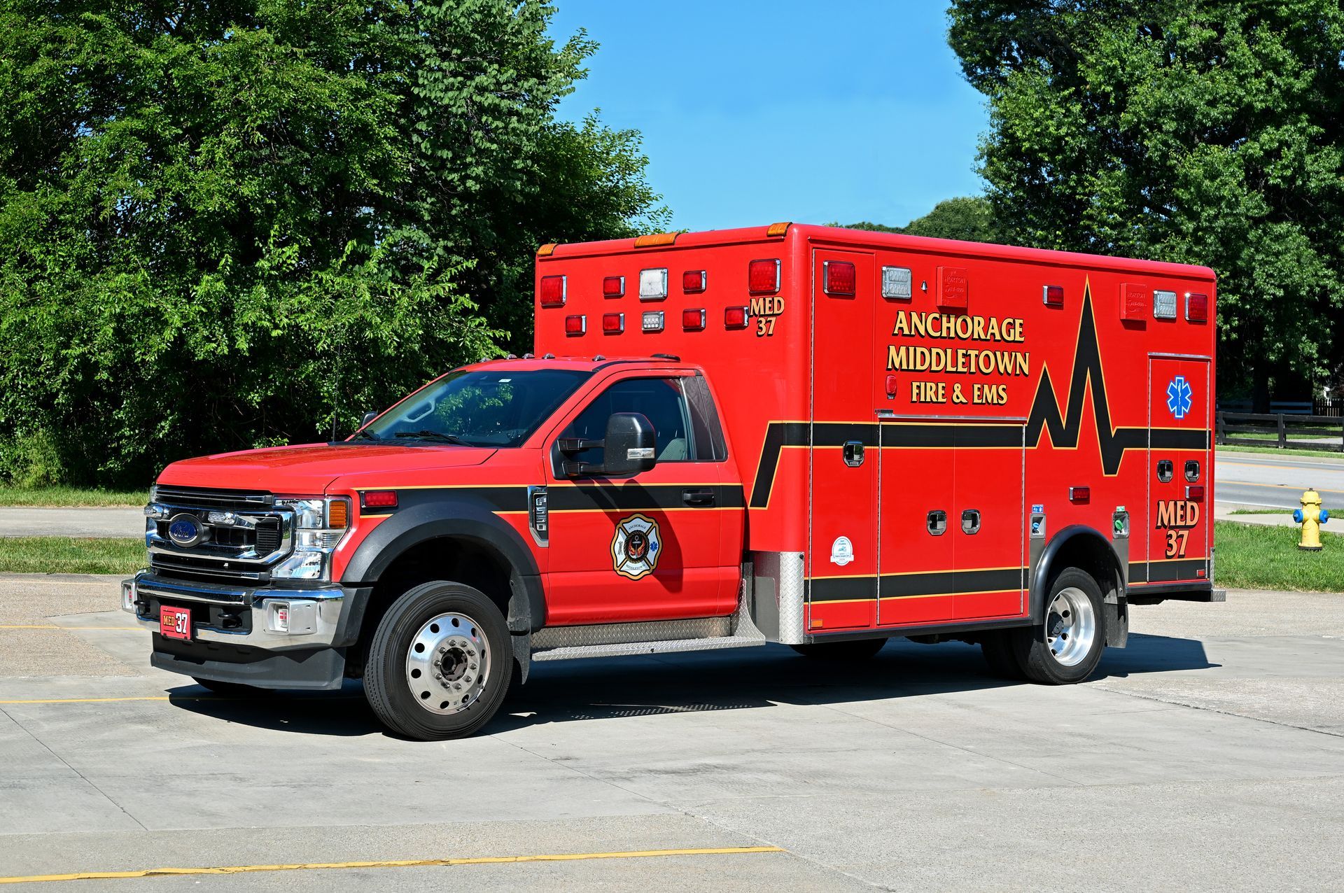 Red ambulance with black trim, parked on asphalt; trees in background. Text on the side says 