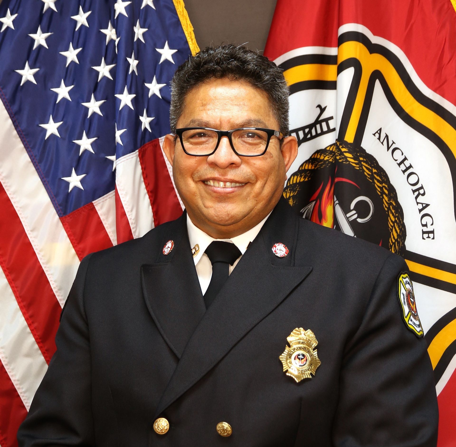 Fire Chief in uniform smiling, with US flag and Anchorage Fire Department flag in background.