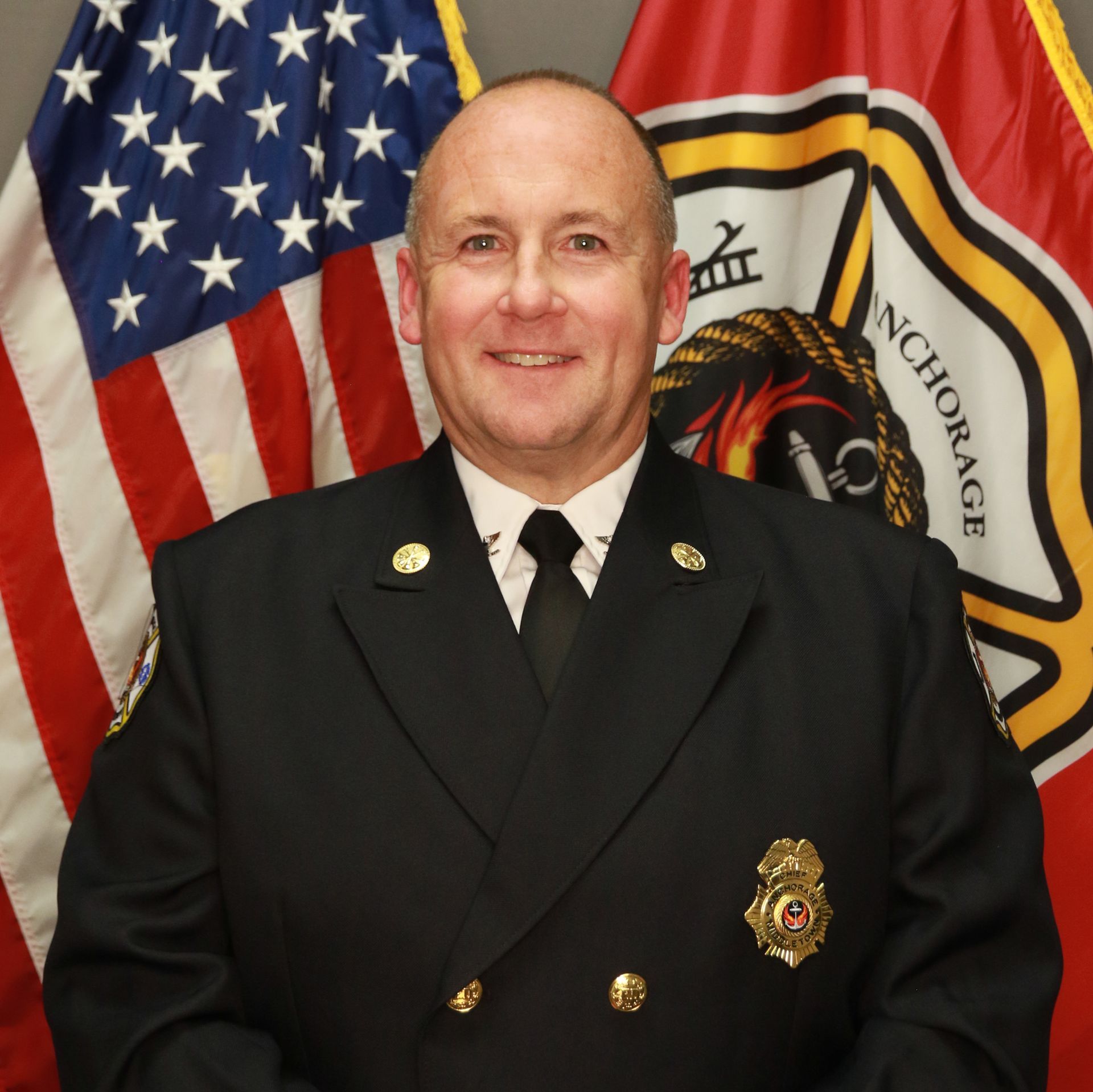 Fire Chief in uniform, smiling in front of US and Anchorage fire department flags.