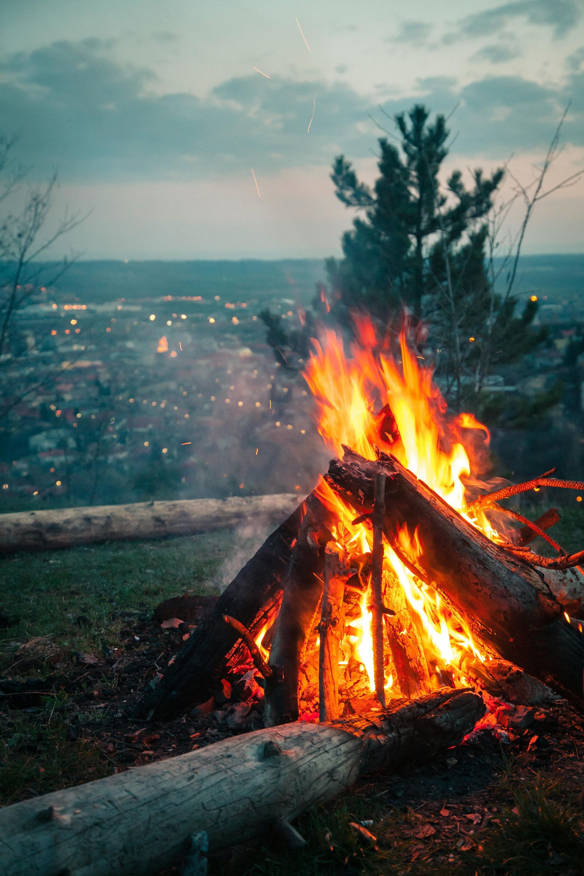 Campfire burning outdoors at dusk with a cityscape in the background.