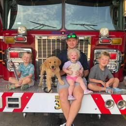 Man, three children, and dog on a fire truck. The man holds a toddler; the other two sit beside them.
