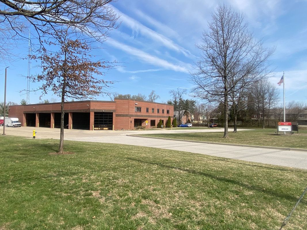 Brick fire station with open bay doors under a blue sky, grass in the foreground.