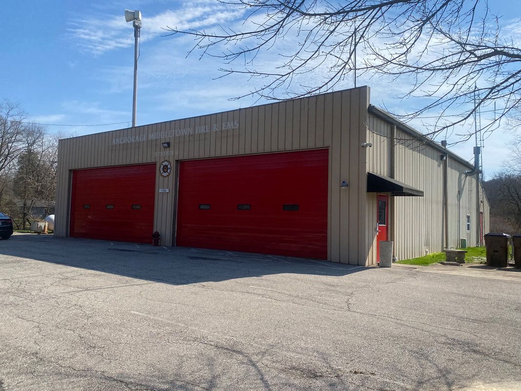 Fire station with red garage doors, beige siding, gravel driveway, and a blue sky.