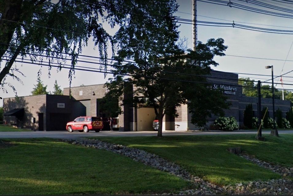 Brick building with a red fire truck parked in front. Green grass and trees are in the foreground.