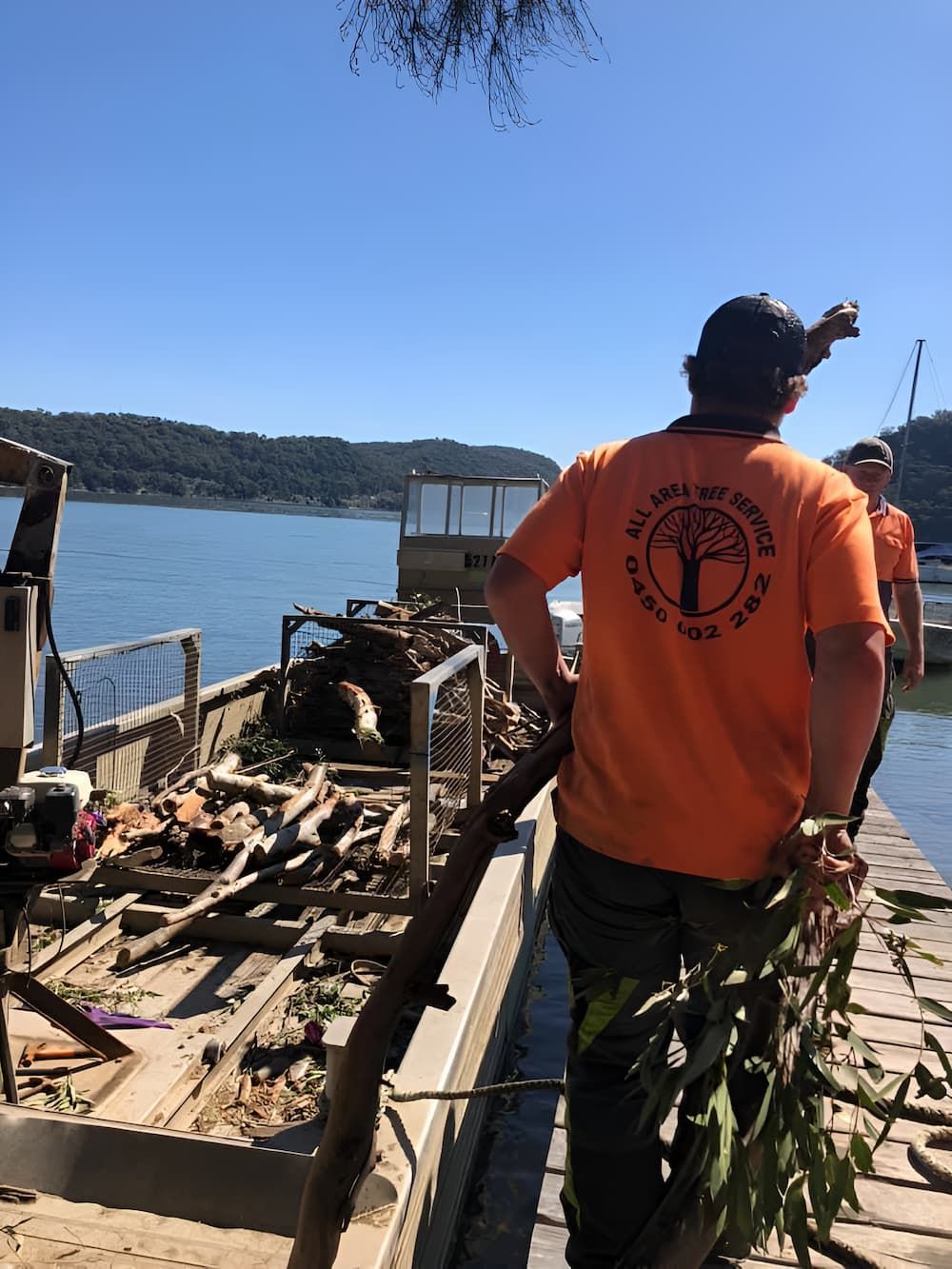 A Man In An Orange Shirt Is Standing On A Dock Next To A Body Of Water — All Area Tree Service Pty Ltd In Fountaindale, NSW