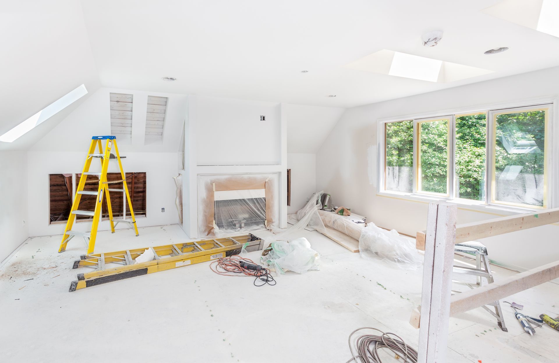 Bright white room under renovation with ladders, tools, plastic-covered furniture, and large windows Bright white room under renovation with ladders, tools, plastic-covered furniture, and large windows