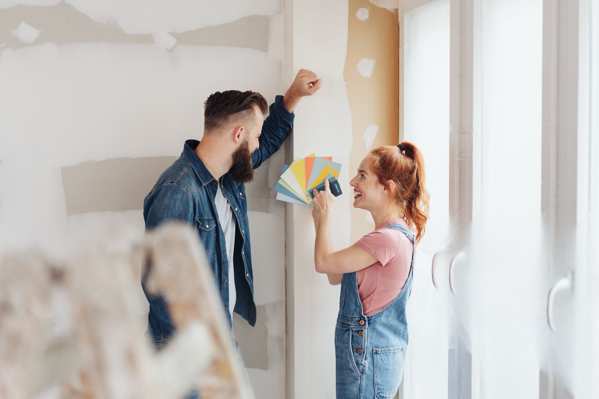 Two people paint a room, with one holding color swatches near a partially finished wall. Two people paint a room, with one holding color swatches near a partially finished wall.