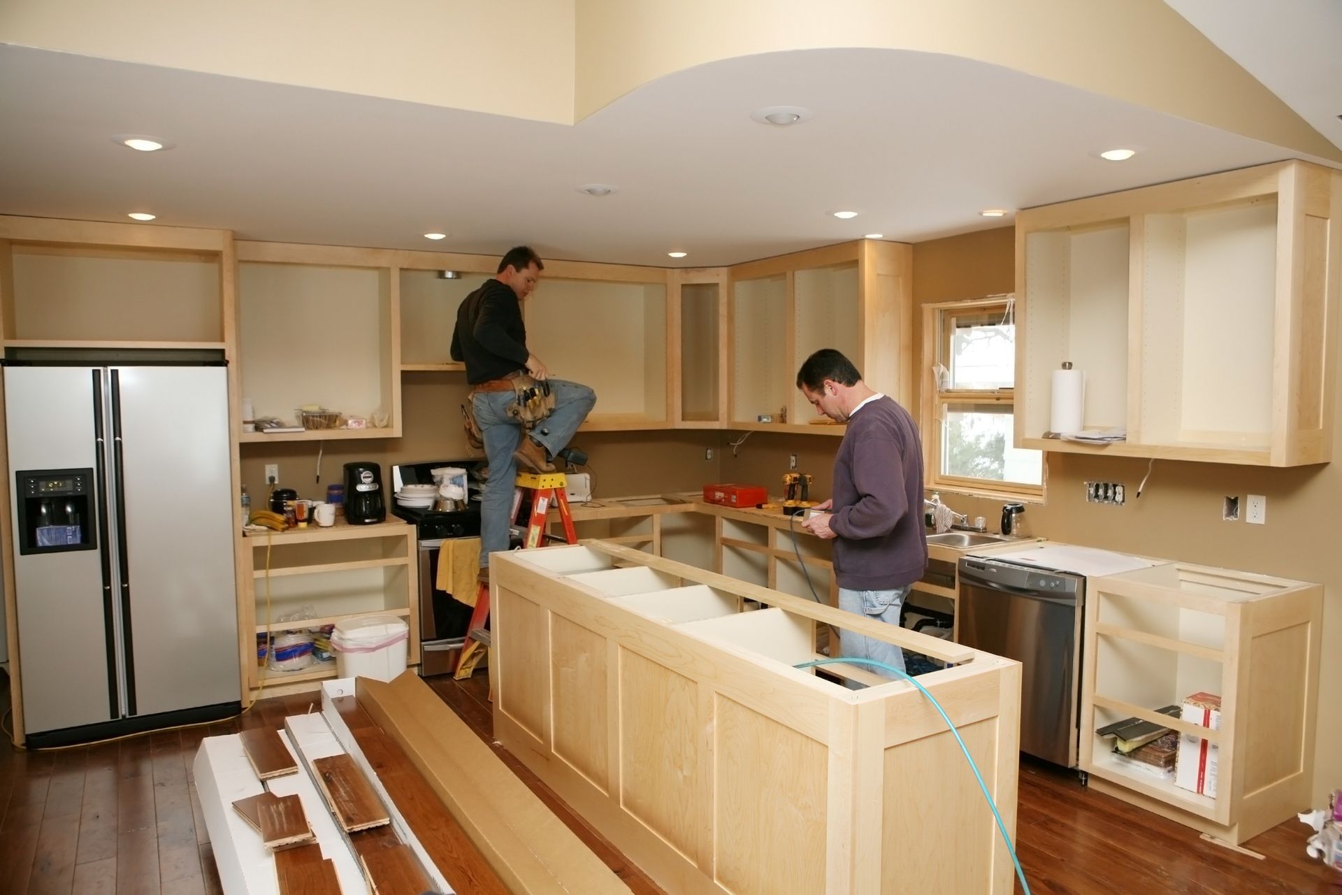 Two people installing custom wooden cabinetry in a kitchen under renovation with a stainless steel refrigerator.