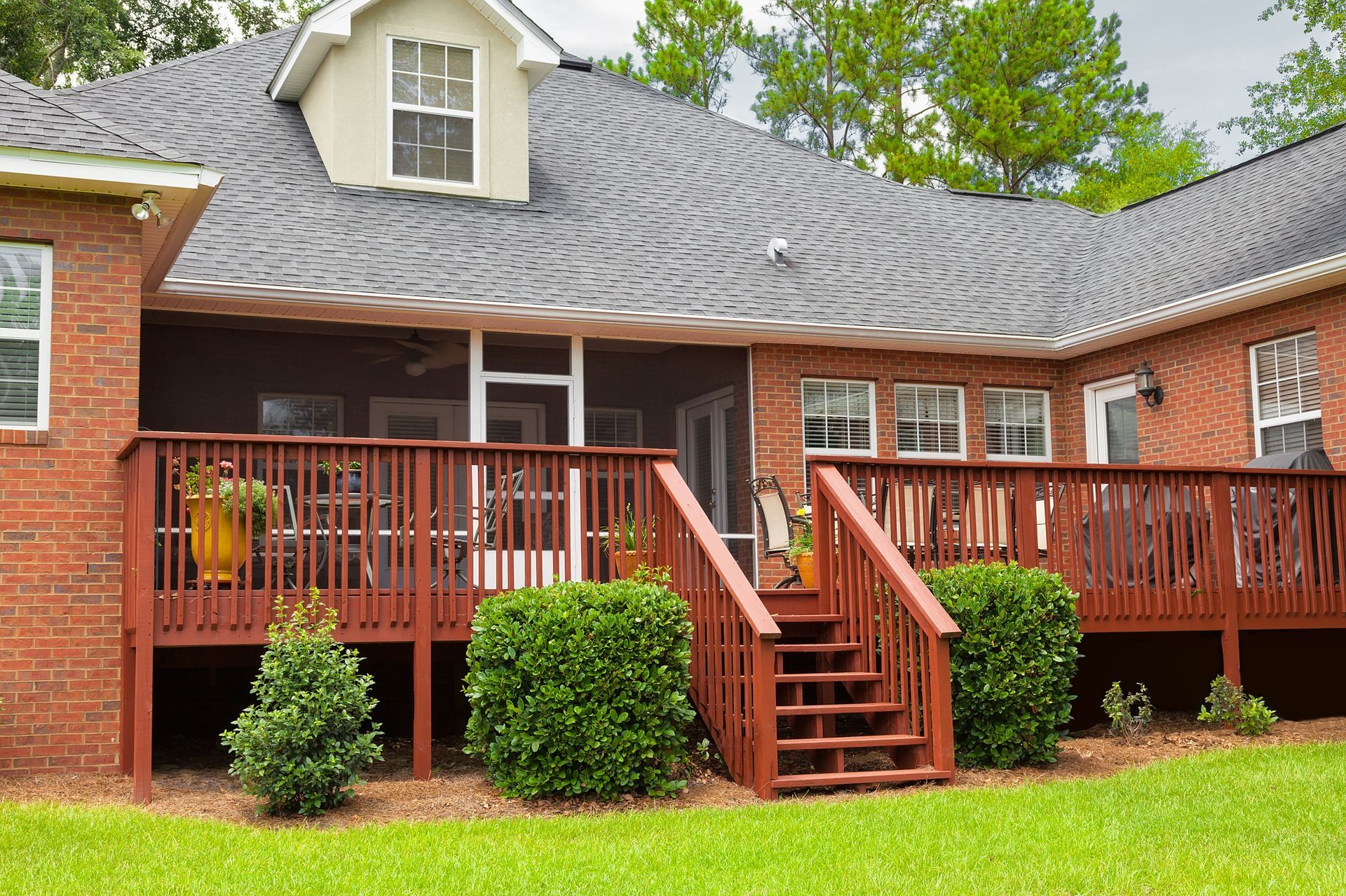 A red-brick house featuring a large, wooden deck with stairs and a screened-in porch, set against a green lawn.