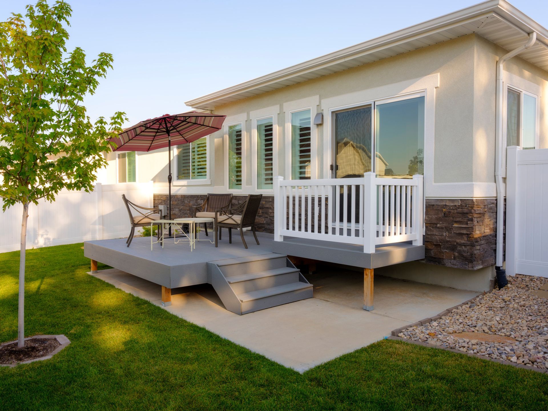 A tan house with a gray deck, white railing, and patio furniture under a red umbrella, set in a sunny backyard.
