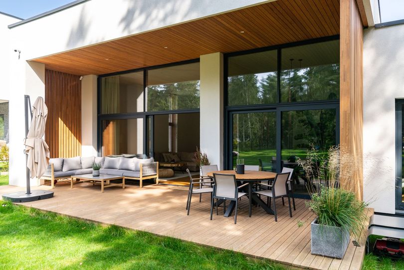 A modern patio with light wood decking, outdoor lounge seating, and a dining set under a covered roof in a green yard.