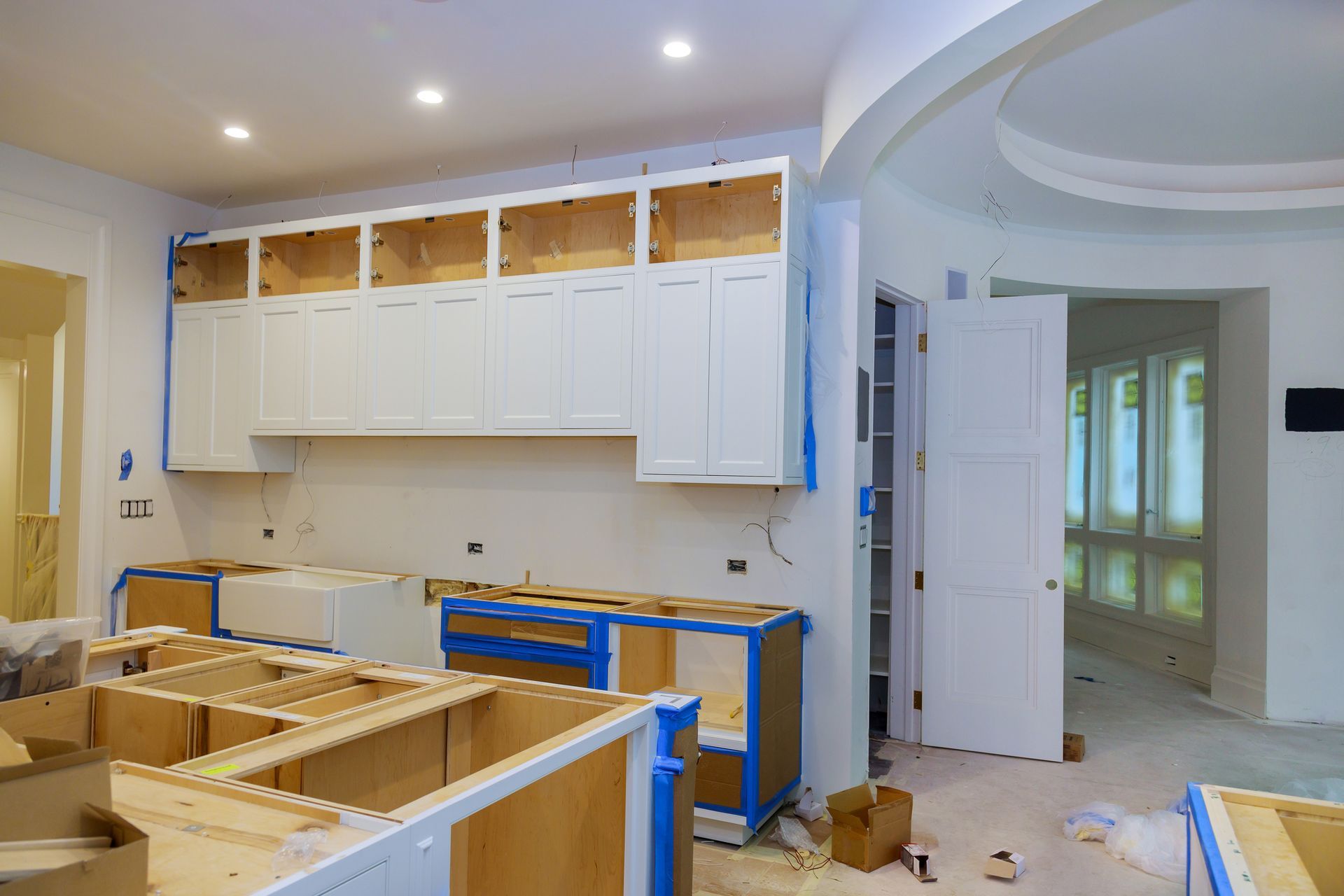A kitchen under construction with white upper cabinets and base cabinets with blue painters tape in a bright room.