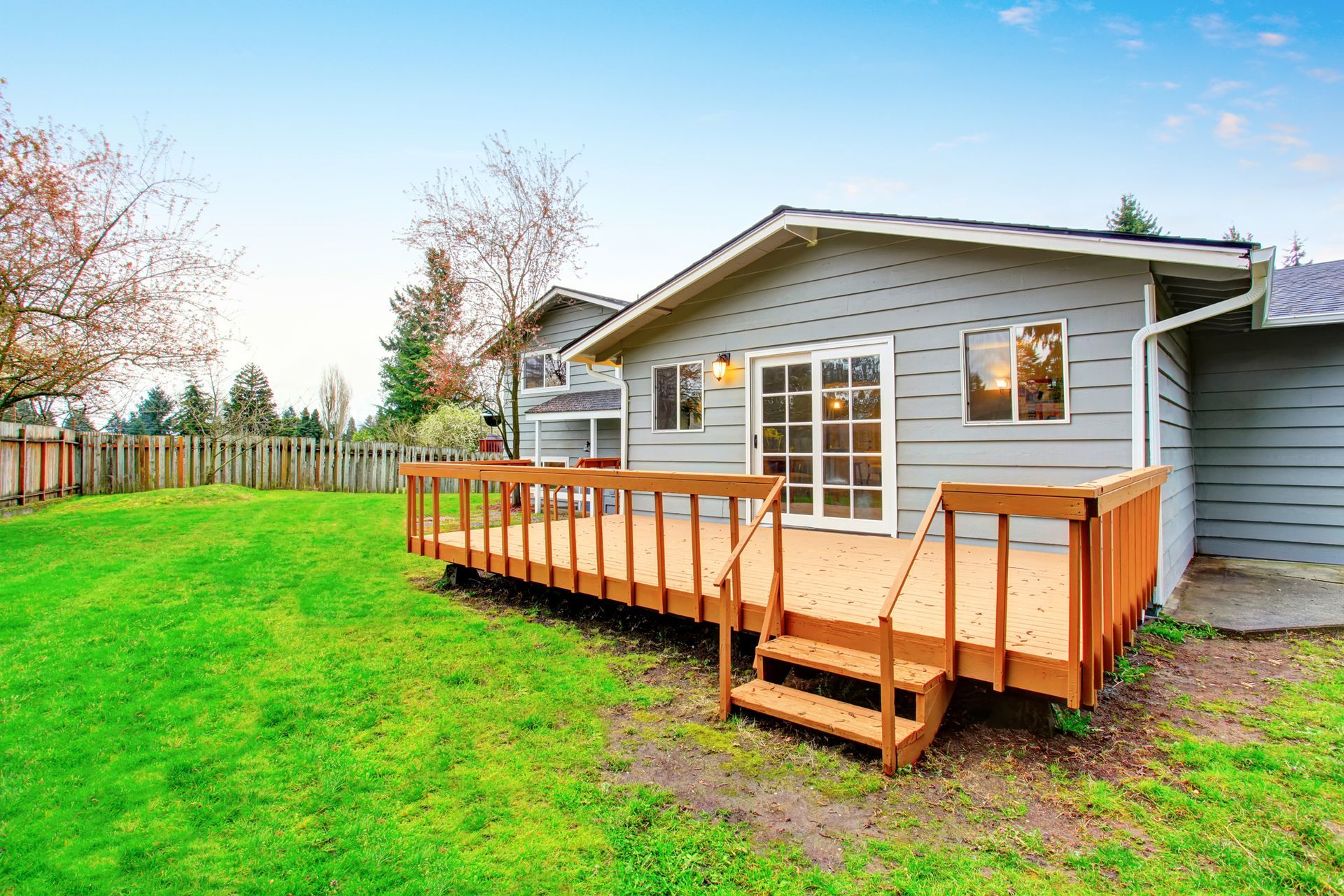 A light grey, single-story house with a wooden deck and sliding glass doors, overlooking a large green backyard.