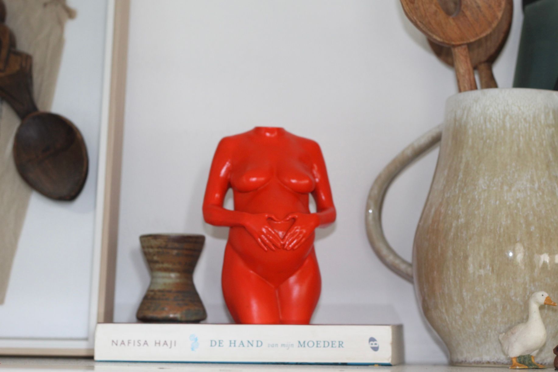 Een rood, torso-vormig beeld op een boekenplank naast een grote beige vaas en hangende houten gebruiksvoorwerpen.