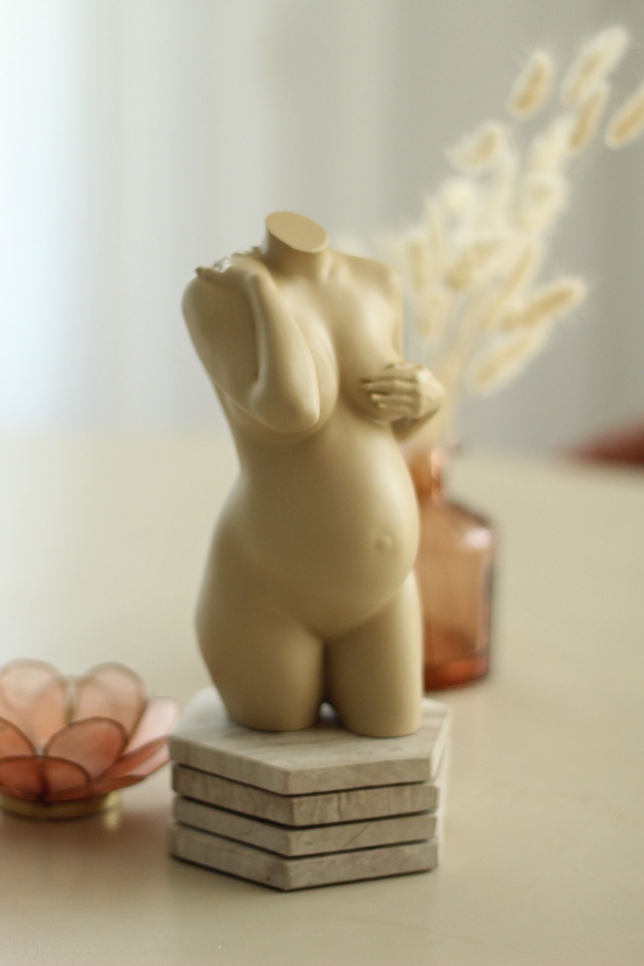 Beige torso-beeldje op een stapel boeken, met gedroogde bloemen in een zacht verlichte kamer.