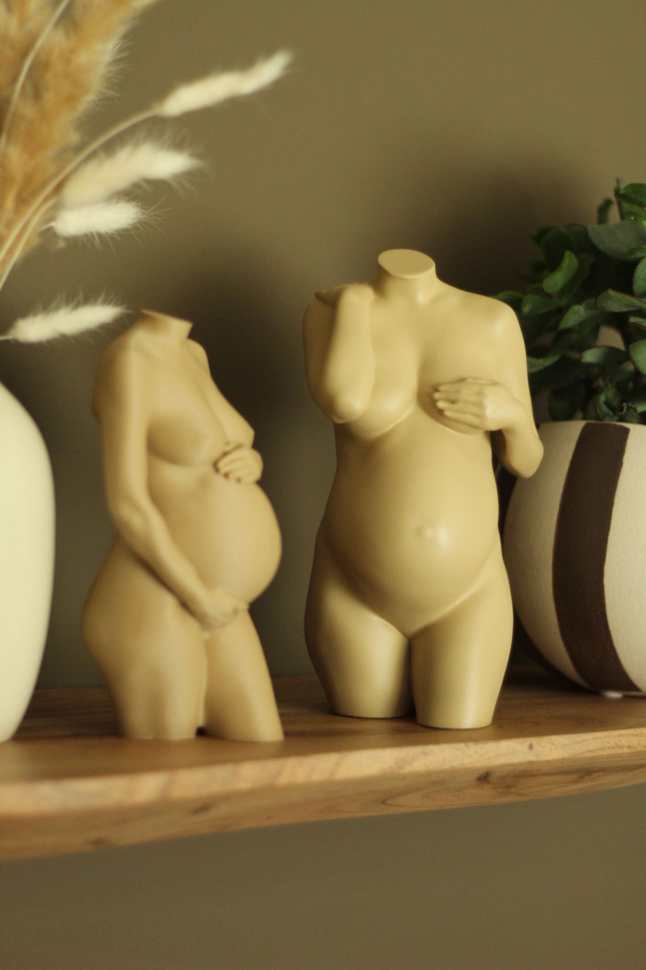 Twee crèmekleurige torso-beeldjes op een houten plank, met een vaas en groen op de achtergrond.