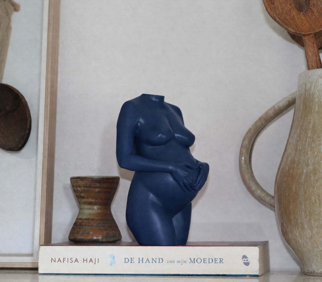 Een blauwe sculptuur van een torso zonder hoofd, die zijn buik vasthoudt, staat op een witte plank, geflankeerd door geweven decoratieve stukken.
