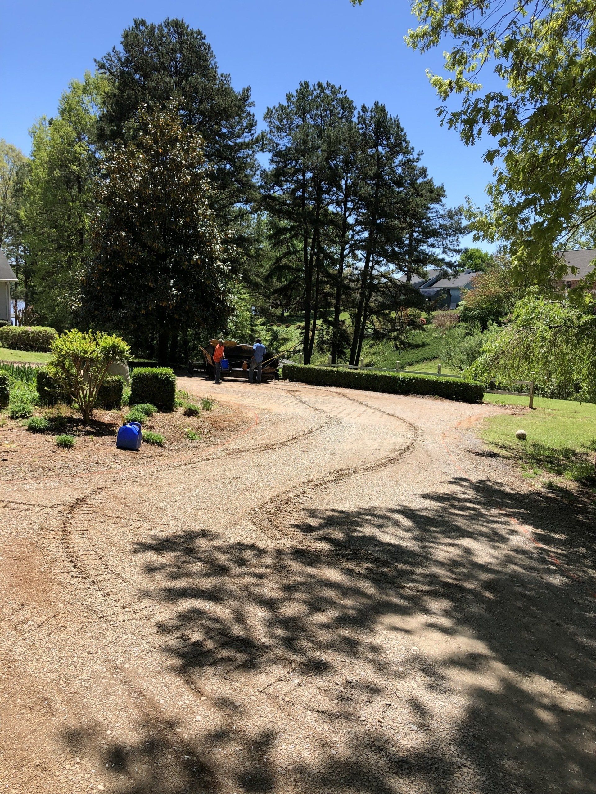 Gravel driveway with a tractor, trees, and blue bucket under a sunny sky.