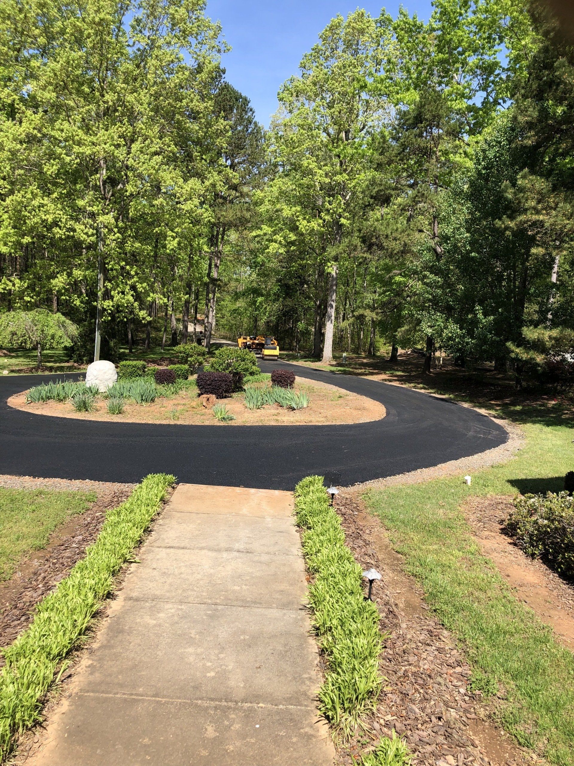 Asphalt circular driveway with central flowerbeds, accessed via a concrete walkway lined with hedges. Trees surround.