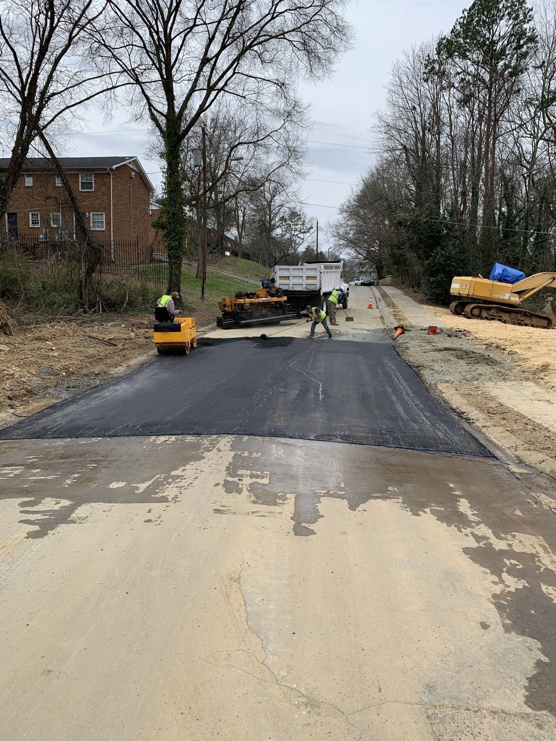 Road construction: workers paving a street with fresh asphalt. Equipment and trees in the background.