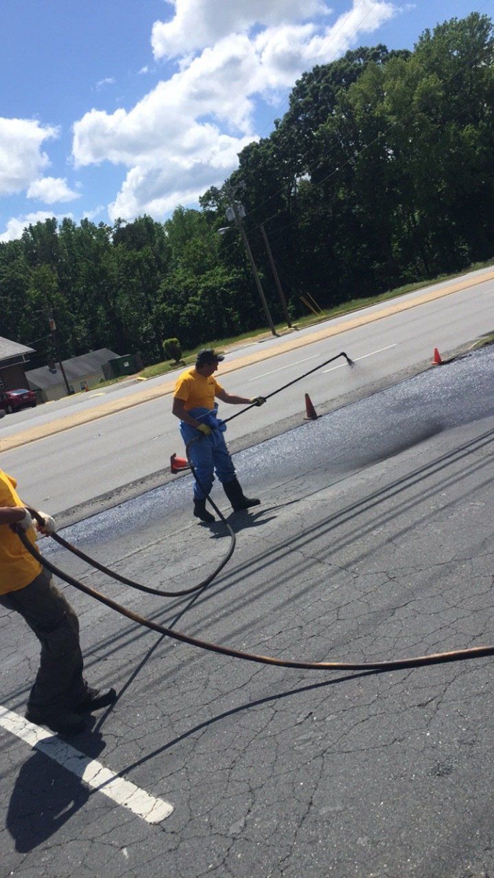 Two workers in yellow shirts spraying asphalt on a road, under a blue sky.