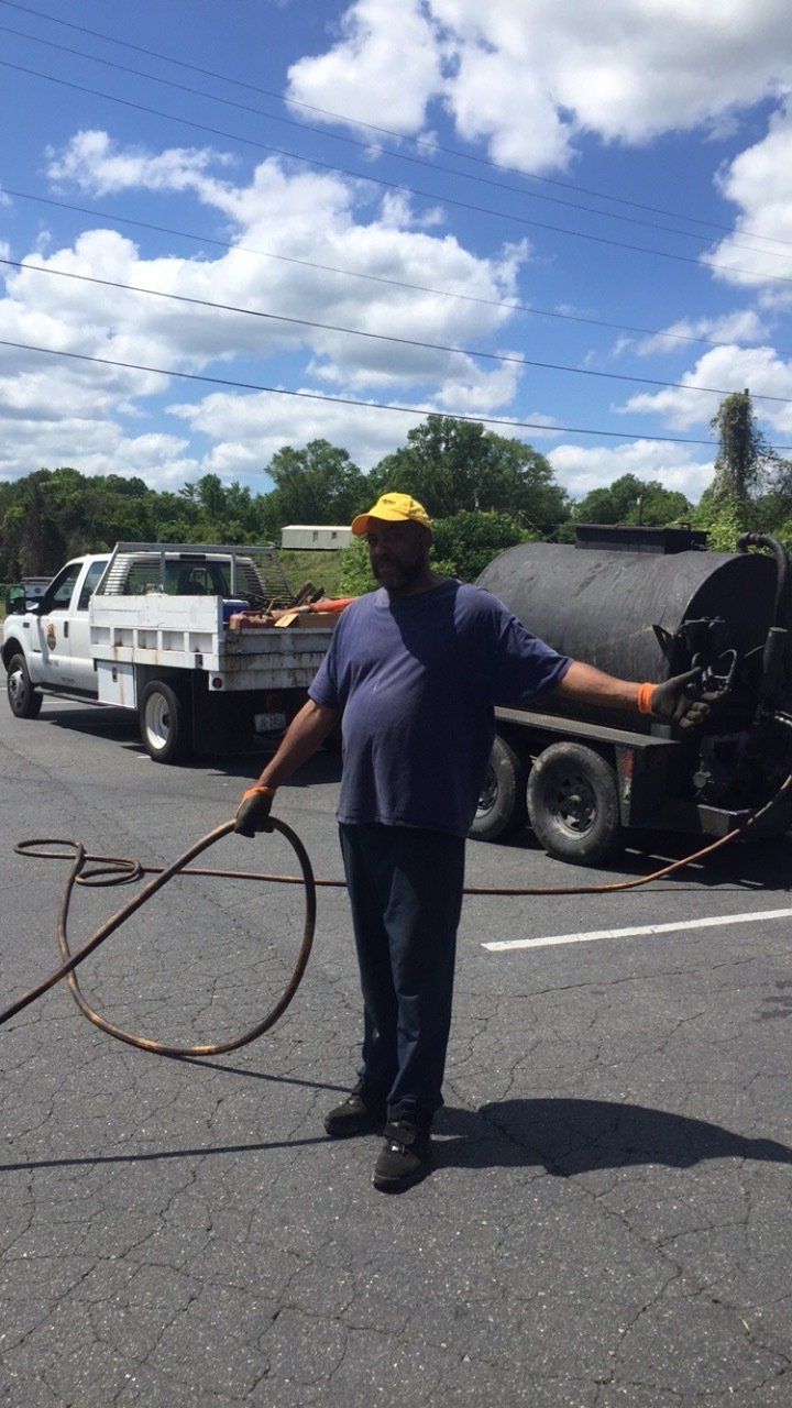 Man in yellow hat and blue shirt holding a hose near a truck and asphalt tank. Sunny outdoor setting.