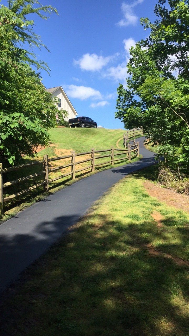 Asphalt driveway leading uphill, framed by a wooden fence and grass, toward a house under a sunny sky.