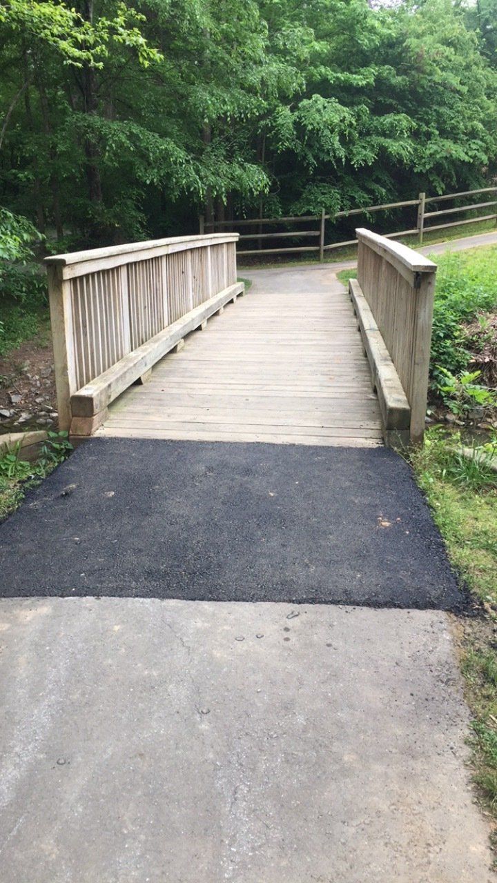 Wooden bridge over a small creek in a park; asphalt path leads to the bridge.