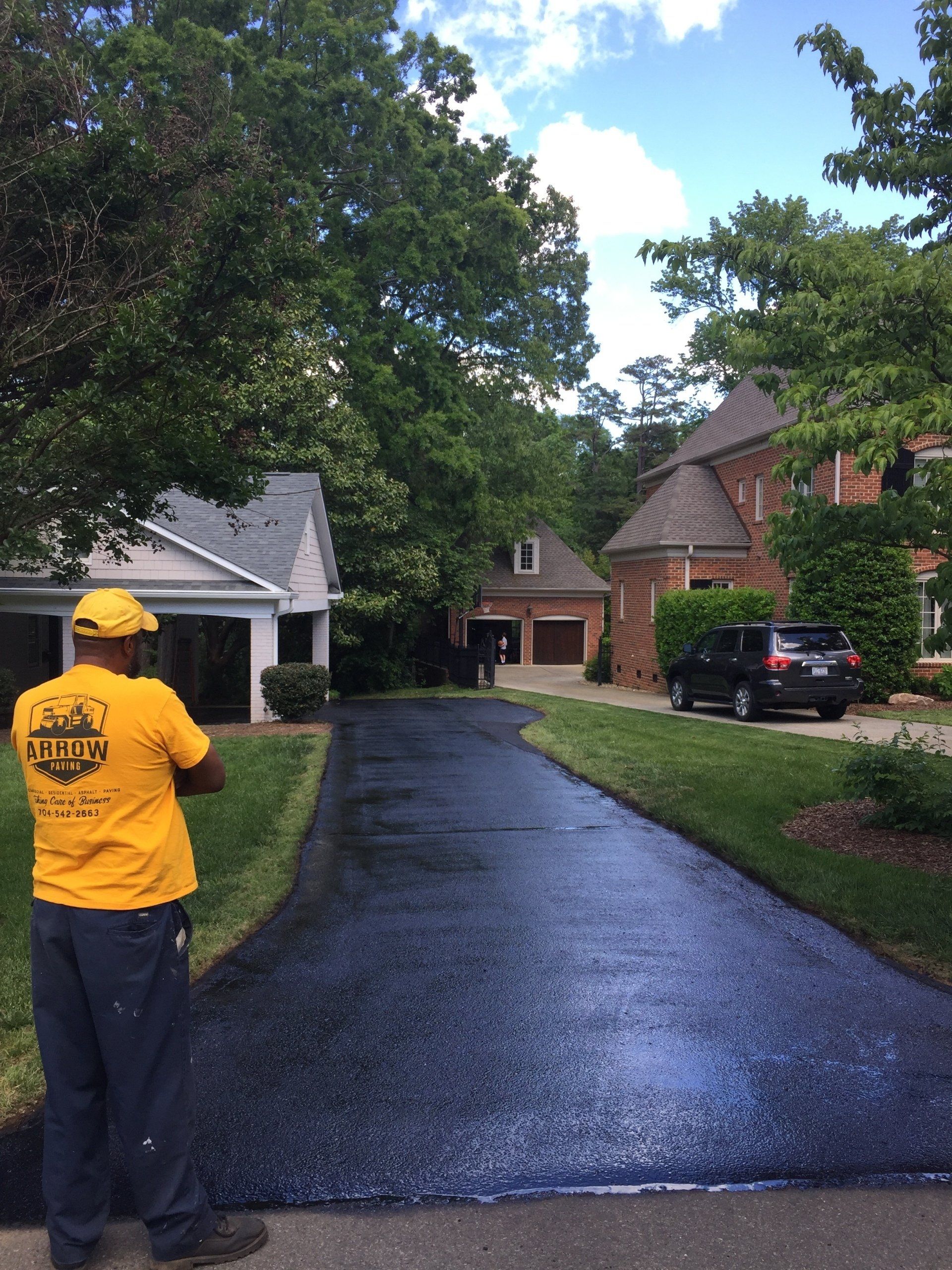 Person in yellow shirt stands beside a newly paved asphalt driveway in front of houses with trees and blue sky.