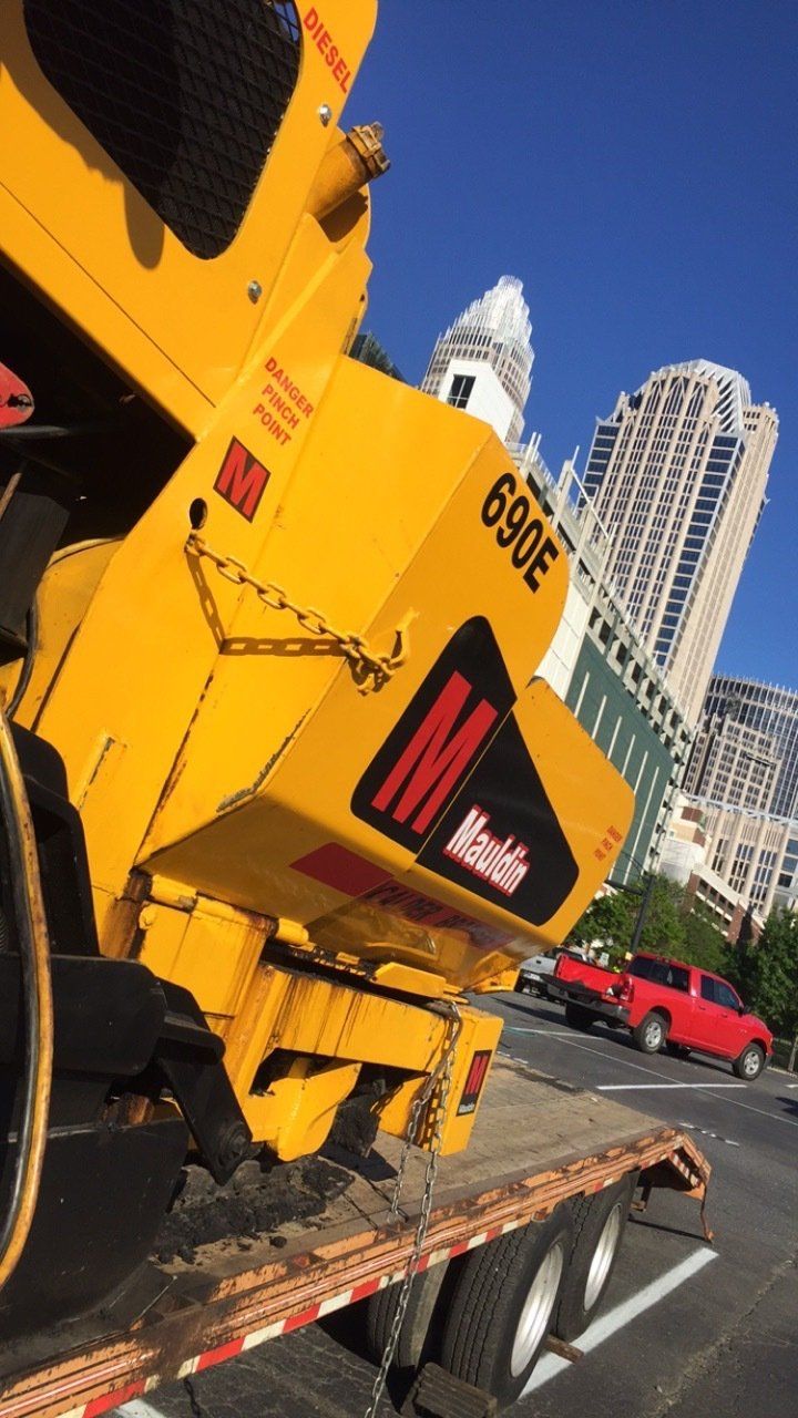 Yellow street sweeper on a flatbed truck in an urban setting; red vehicle in background.
