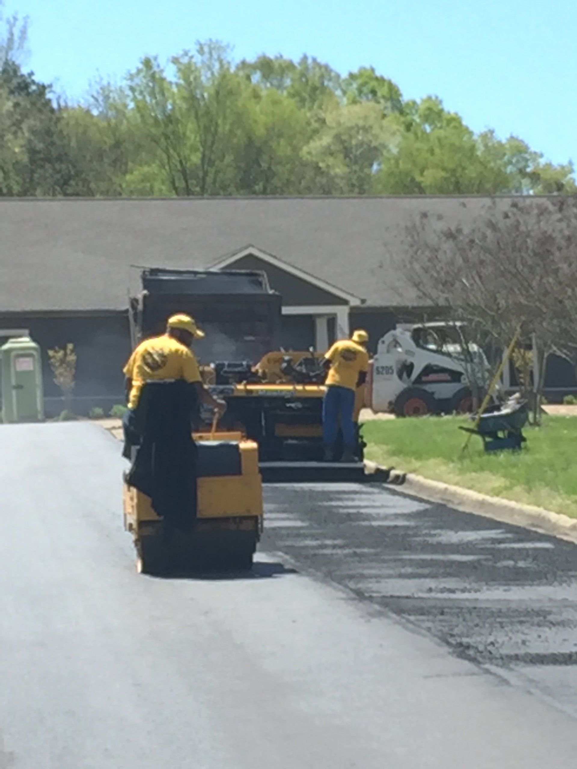 Road paving crew with asphalt roller, paving equipment, and skid steer loader on a road in front of a building.