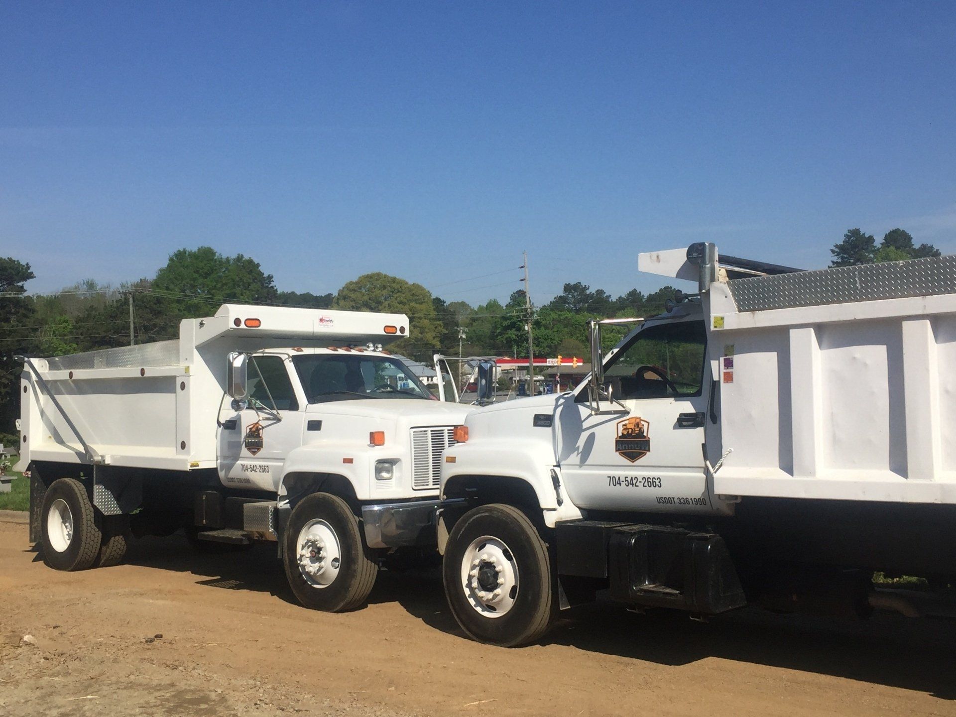 Two white dump trucks parked on dirt under a blue sky, trees in the background.
