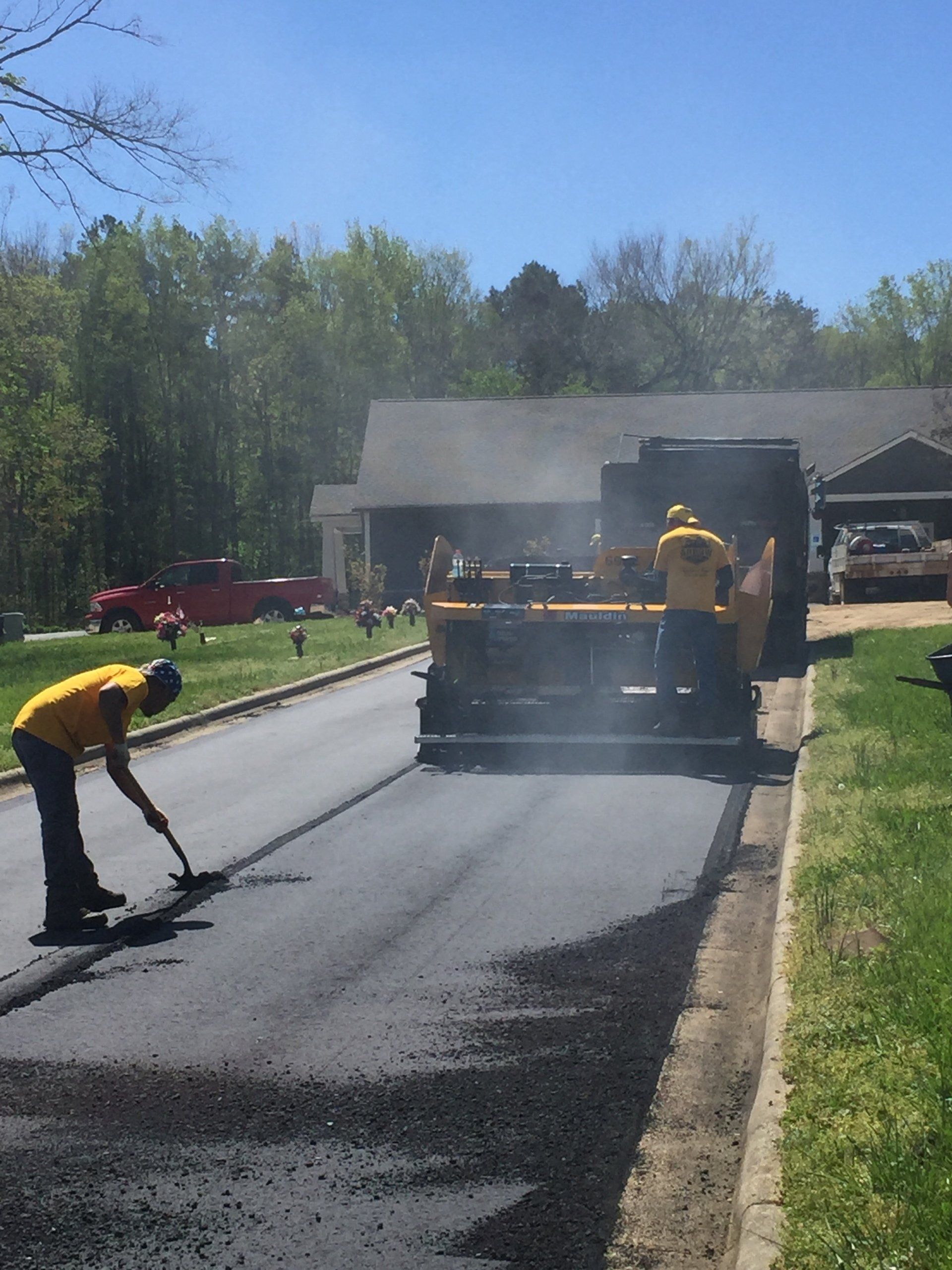 Asphalt paving crew laying fresh pavement on a residential street. Yellow-shirted workers operate machinery and hand tools.